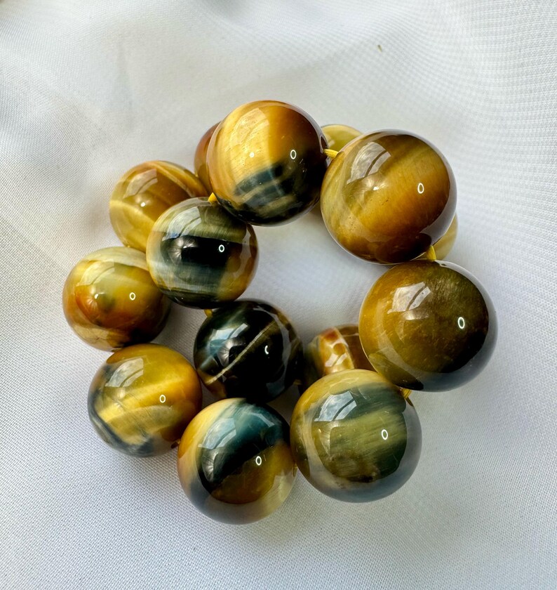 May include: A bracelet featuring large, polished tiger's eye beads. The beads display a mix of golden brown and deep blue-green hues, strung together on a yellow cord. The spherical beads have a glossy finish.