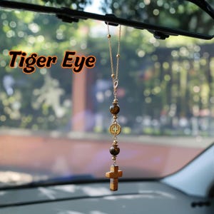 May include: A car rearview mirror hanging ornament featuring a gold-toned chain with two round brown tiger eye beads, a small circular medal with a cross emblem, and a carved tiger eye cross pendant at the bottom. The text Tiger Eye is visible.