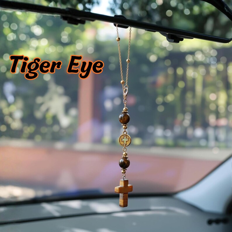 May include: A car rearview mirror hanging ornament featuring a gold-toned chain with two round brown tiger eye beads, a small circular medal with a cross emblem, and a carved tiger eye cross pendant at the bottom. The text Tiger Eye is visible.