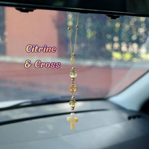 May include: A gold-toned chain car accessory featuring two translucent yellow citrine beads, a circular metal charm with a cross cutout, and a light-colored stone cross pendant at the bottom. The text Citrine & Cross is visible on the left.