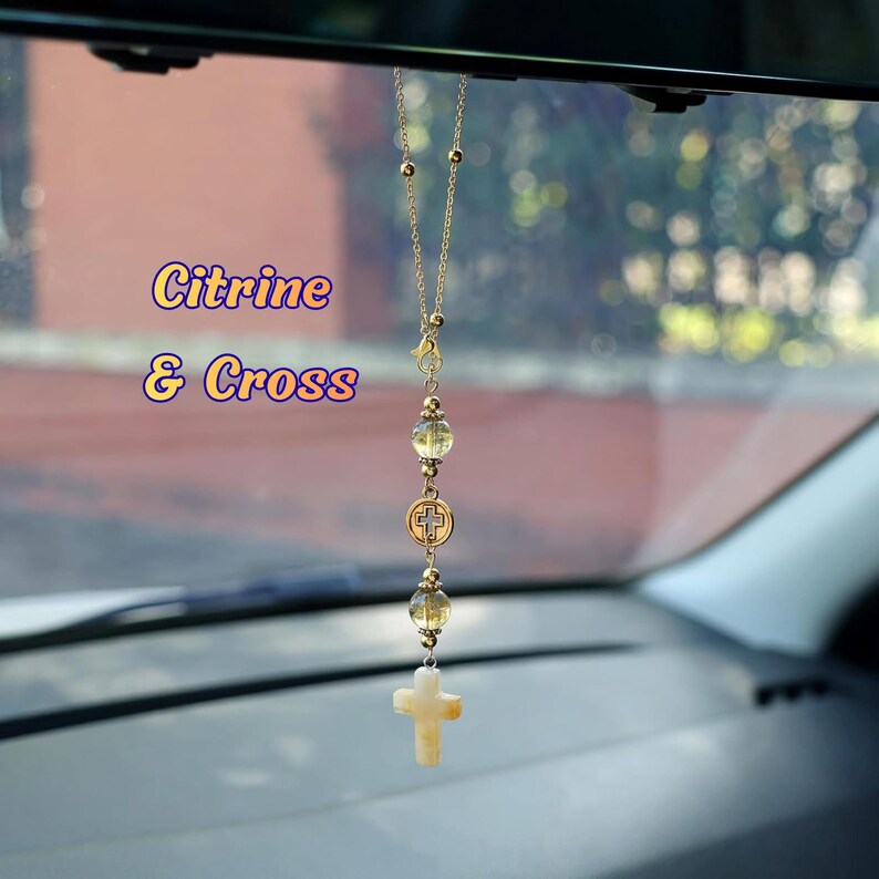 May include: A gold-toned chain car accessory featuring two translucent yellow citrine beads, a circular metal charm with a cross cutout, and a light-colored stone cross pendant at the bottom. The text Citrine & Cross is visible on the left.