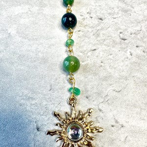 May include: A gold-toned sun pendant featuring a central iridescent cabochon, suspended from a gold-toned ball chain adorned with a vertical arrangement of five green beads in varying shades and sizes, connected by small gold-toned metal jump rings.