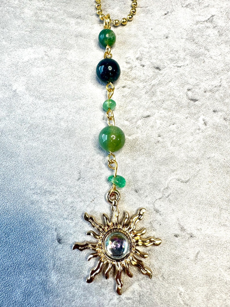 May include: A gold-toned sun pendant featuring a central iridescent cabochon, suspended from a gold-toned ball chain adorned with a vertical arrangement of five green beads in varying shades and sizes, connected by small gold-toned metal jump rings.