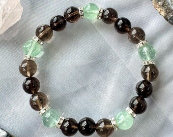 May include: A beaded bracelet featuring alternating smoky quartz and green fluorite gemstone beads. The bracelet is accented with silver-toned spacers. The gemstones are round and polished, creating a natural and elegant accessory.