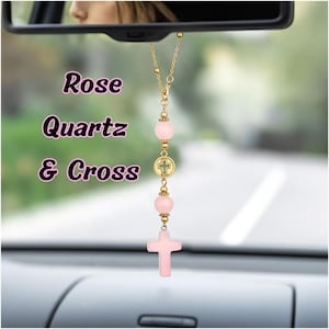 May include: A car rearview mirror hanging ornament featuring a gold-tone chain with two round, pale pink rose quartz beads, a small circular gold-tone charm with a cross emblem, and a larger, carved pale pink rose quartz cross pendant at the base.