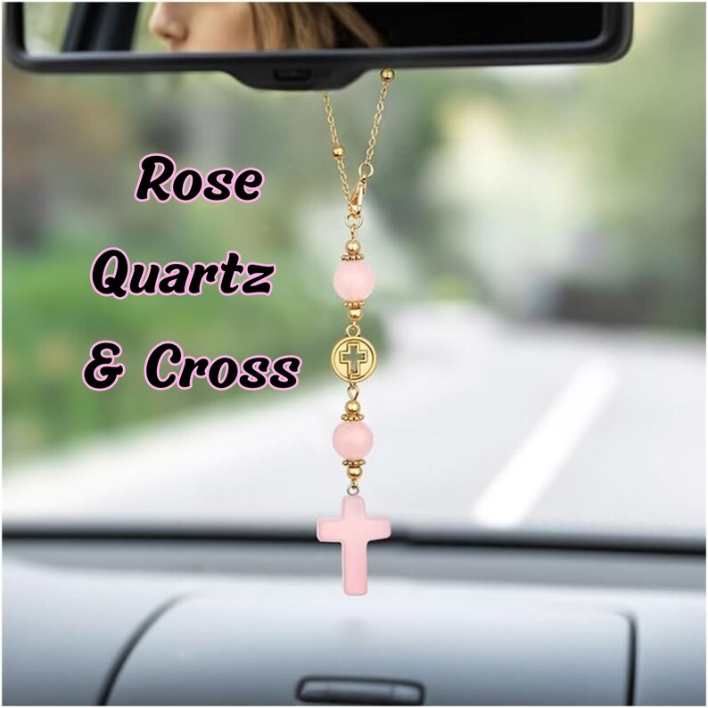 May include: A car rearview mirror hanging ornament featuring a gold-tone chain with two round, pale pink rose quartz beads, a small circular gold-tone charm with a cross emblem, and a larger, carved pale pink rose quartz cross pendant at the base.