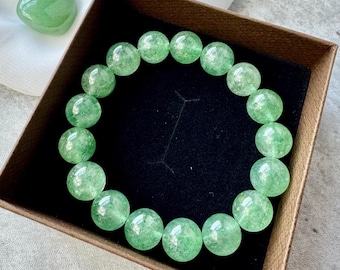 May include: A green beaded bracelet with large, round beads. The bracelet is displayed in a brown box with a black interior. A green, polished stone sits next to the bracelet.