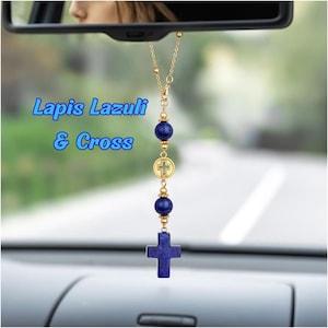 May include: A car rearview mirror hanging ornament featuring a gold-tone chain, two round deep blue lapis lazuli beads, a small gold-tone circular charm with a cross cutout, and a larger lapis lazuli cross pendant. The text Lapis Lazuli & Cross is visible.