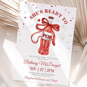 May include: A baby shower invitation with a red and white design. The invitation features a watercolor illustration of a champagne bottle with a red bow and the word "POP". The text reads "She's Ready To" and includes the mom-to-be's name, date, and time.