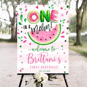 Puede incluir: Un letrero decorativo con un tema de sandía. El letrero dice "ONE in a Melon!" y "Welcome to Brittani's First Birthday!" en letras rosas y verdes. El fondo tiene rayas rosas y blancas con corazones.