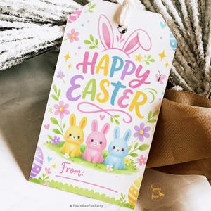 May include: A rectangular gift tag with the words "Happy Easter" in colourful letters, featuring bunny ears and three cartoon bunnies. The tag includes space to write "From:" and is decorated with flowers and eggs.