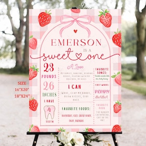 Op de afbeelding: Een roze en rood verjaardagsbord met de tekst "Emerson is a sweet one". Het bord bevat het gewicht, de lengte en de favoriete dingen van het kind. Het bord is versierd met aardbeien en een roze strik.