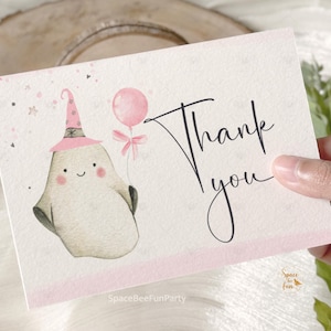 Może przedstawiać: Prostokątna kartka okolicznościowa z jasnobeżowym duszkiem w różowym czapeczce imprezowej, trzymającym różowy balonik. Napis Thank you wykonany czarną kursywą znajduje się po prawej stronie. Tło jest białe z różowymi akcentami.