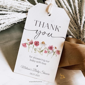 May include: A white gift tag with the words "THANK you" in elegant script, adorned with watercolor flowers. The tag reads "for showering our baby with love" and "Melissa's Baby Shower 08.27.2021". A small logo is at the bottom.