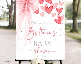 Könnte beinhalten: Ein weißes Willkommensschild für eine Babyparty auf einer schwarzen Staffelei. Das Schild zeigt eine große rosa Schleife, rosa Herzen und den Text "Welcome to Brittani's BABY shower" mit dem Datum 11.27.2020. Ein Strauß weißer Blumen befindet sich am Fuß.