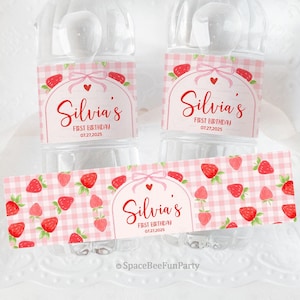 Op de afbeelding: Waterflesetiketten met een roze ruitpatroon en aardbeienillustraties. De etiketten tonen de naam "Silvia's" in rood script, met "FIRST BIRTHDAY" en de datum "07.27.2025". Een roze strik en hart accentueren het ontwerp.