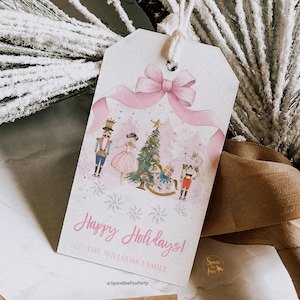 May include: A white gift tag with a pink bow and ribbon design. The tag features a watercolor illustration of a Christmas tree, nutcrackers, and a mouse. The text "Happy Holidays!" is written in pink.