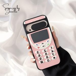 May include: A pink phone case with a retro design, featuring a keypad and screen. The case has a black border and white accents, with the word "Nokia" printed on the top. The case is held in a hand.