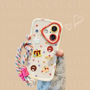 Puede incluir: Funda transparente para teléfono con pegatinas temáticas de caramelos y un anillo rojo para la cámara. Cuenta con una correa de muñeca arcoíris. El texto "VIOLETA CREATES" es visible.