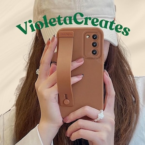 Puede incluir: Una funda de teléfono marrón con una correa a juego, con tres lentes de cámara. La funda tiene el texto "zaya lagos" y la sostiene una persona. El texto "Violeta Creates" está en verde sobre el teléfono.