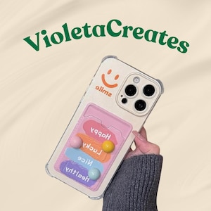 May include: A beige phone case with a pink card holder, featuring a smiley face and the word "olime". The card holder has colorful stripes with the words "Happy", "Lucky", "Nice", and "Health". The text "Violeta Creates" is in green.