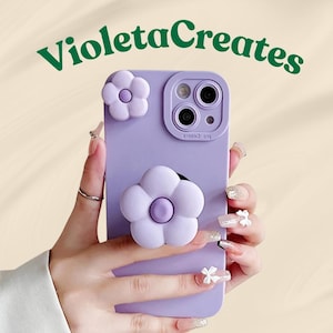 May include: A lilac phone case featuring two white flower decorations. The case has a camera aperture and the text "VioletaCreates" in green script. The phone case is held by a person with manicured nails.