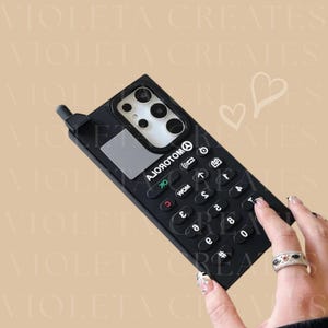 May include: Black phone case with a retro keypad design. The case has a rectangular shape with a small antenna and a screen area. The keypad features white numbers and symbols. The text "AJОЯОТОМО" is printed on the case.
