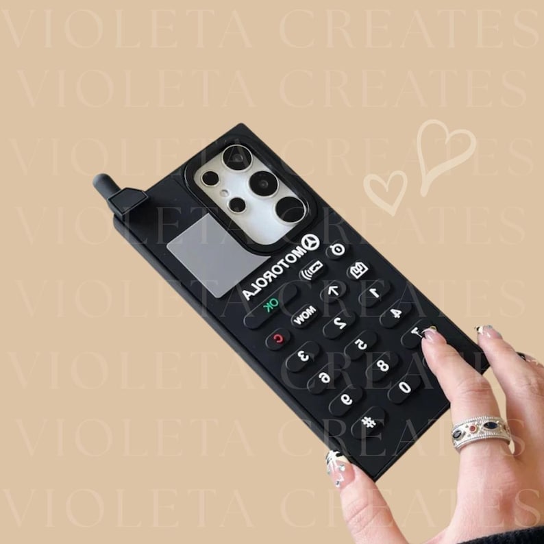May include: Black phone case with a retro keypad design. The case has a rectangular shape with a small antenna and a screen area. The keypad features white numbers and symbols. The text "AJОЯОТОМО" is printed on the case.