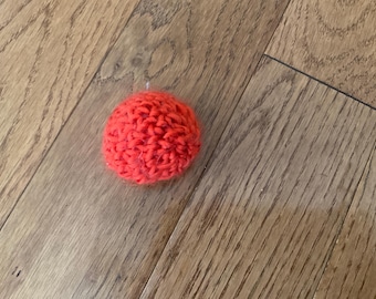 May include: A bright red, round, knitted pom-pom, possibly for a hat or other craft project. The pom-pom is made of a textured yarn and is resting on a wooden surface.