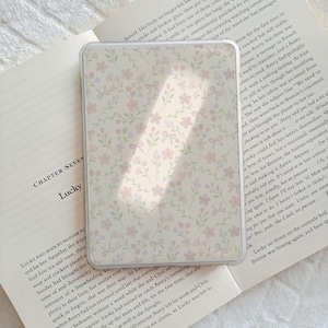 May include: A transparent phone case featuring a floral design with pink flowers and green leaves. The case is placed on an open book, creating a soft, feminine aesthetic. The design is simple and elegant, with a light colour palette.