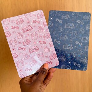 Può includere: Due biglietti rettangolari a tema libro. Un biglietto è rosa con illustrazioni bianche di libri, occhiali, cuffie e tazze di caffè. L'altro biglietto è blu navy con illustrazioni bianche simili. I biglietti sono tenuti da una persona.