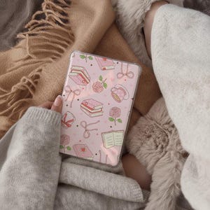 May include: A pink phone case featuring a pattern of books, roses, bows, and teacups. The case is held in a person's hand, with a cozy blanket and slippers in the background. The overall aesthetic is soft and feminine.