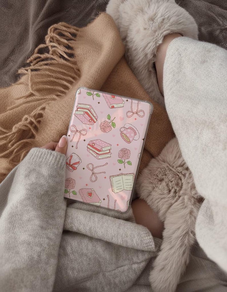 May include: A pink phone case featuring a pattern of books, roses, bows, and teacups. The case is held in a person's hand, with a cozy blanket and slippers in the background. The overall aesthetic is soft and feminine.