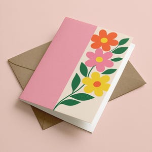 May include: A pink greeting card with a floral design featuring orange, pink, and yellow flowers with green leaves. The card is paired with a brown envelope, all set against a soft pink background. The card is a perfect gift for any occasion.