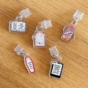 May include: Five clear acrylic keychains with various book-themed designs. One keychain features an open book with the text "In a really good place right now." Another says "In my book of course not mentally." Other designs include a coffin, matches, and a Kindle.