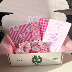 Peut inclure: Une boîte cadeau rose remplie de papeterie et d'accessoires. Comprend un carnet, des autocollants, un stylo et un chouchou. La boîte contient une carte de bibliothèque avec le texte "Buy me books and tell me I'm pretty". La boîte est étiquetée "70% Recycled".
