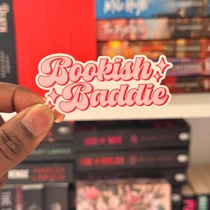 May include: A pink and white sticker with the words "Bookish Baddie" in a retro font. The sticker has a white border and star accents. The background features a bookshelf with various books.