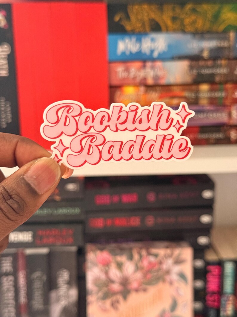 May include: A pink and white sticker with the words "Bookish Baddie" in a retro font. The sticker has a white border and star accents. The background features a bookshelf with various books.