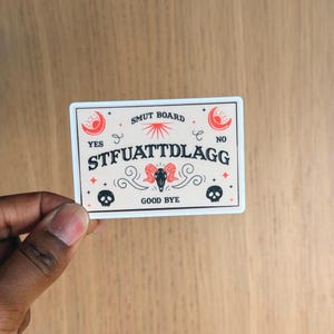 Op de afbeelding: Een rechthoekige sticker met een beige achtergrond en zwarte tekst met de tekst "SMUT BOARD". De sticker heeft een decoratief ontwerp met een schedel, halve manen en de tekst "STFUATTDLAGG". De sticker heeft ook de woorden "YES", "NO" en "GOOD BYE".