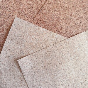 May include: Overhead shot of multiple cork sheets, displaying a natural, speckled pattern. The sheets, in shades of light brown and beige, are arranged at different angles. The cork's surface has a slightly rough texture, emphasizing its organic nature.