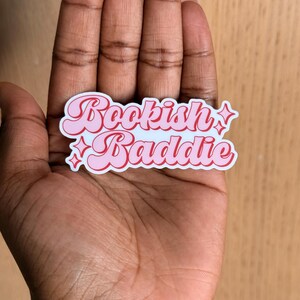 May include: A pink and white sticker with the words "Bookish Baddie" in a retro font. The sticker has a pink border and star accents. The sticker is held in a person's hand.