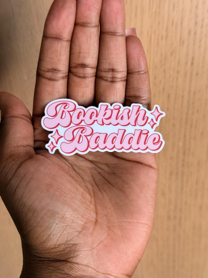 May include: A pink and white sticker with the words "Bookish Baddie" in a retro font. The sticker has a pink border and star accents. The sticker is held in a person's hand.