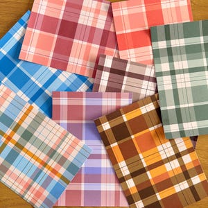 May include: A selection of square paper sheets with plaid patterns in various colours. The sheets feature a mix of colours, including blue, pink, red, green, brown, and orange, with white grid lines creating the plaid design.