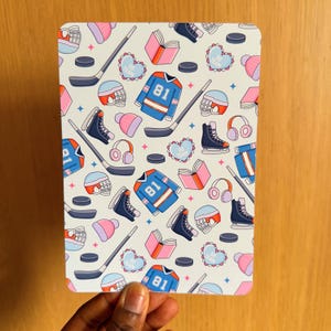 May include: A rectangular, patterned item featuring a repeating design of hockey-related illustrations. The design includes hockey jerseys with the number 81, hockey sticks, skates, helmets, books, and hearts with the words "GO TEAM". The color palette includes blue, pink, and white.