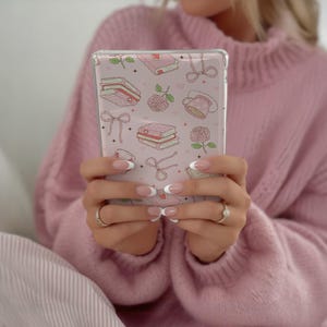 May include: A clear phone case with a pink background and a pattern of pink books, roses, bows, and hearts. The case is held in front of a pink sweater.