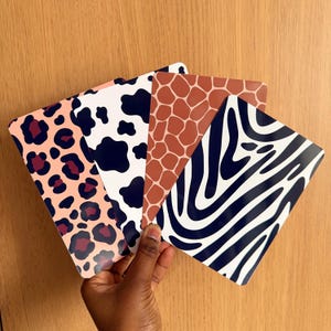 Könnte beinhalten: Eine Hand hält vier rechteckige Karten mit Tiermotiven. Die Muster umfassen Leopardenflecken, Kuhflecken, Giraffenmuster und Zebrastreifen. Die Karten sind fächerförmig vor einem Holzgrund angeordnet.