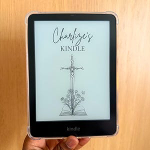 May include: A Kindle e-reader with a clear protective case. The screen displays the text "Charlize's Kindle" above a sword design with roses and butterflies. The word "kindle" is printed at the bottom of the screen.