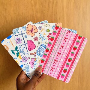 Könnte beinhalten: Ein Set aus fünf rechteckigen Lesezeichen mit abgerundeten Ecken, die verschiedene bunte Muster zeigen, darunter rosa Erdbeeren mit Schleifen, blaue Sardinendosen, tropische Hibiskusblüten und Meeresschildkröten auf weißem Grund.