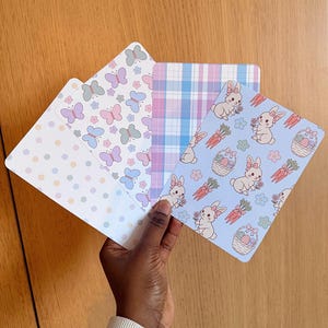 Peut inclure: Une main tient quatre cartes décoratives aux motifs pastel. Les motifs comprennent des papillons, des pois, du tartan et des lapins avec des carottes et des paniers de Pâques. Les cartes sont rectangulaires avec des coins arrondis.