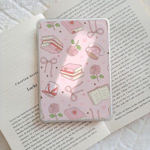 May include: A pink phone case with a repeating pattern of books, roses, bows, and teacups. The design has a soft, feminine aesthetic, perfect for a book lover or someone who enjoys floral motifs.